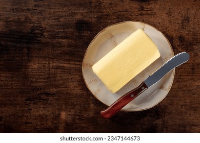 A stick of butter with a knife on a plate, overhead flat lay shot on a dark rustic wooden background with a place for text - Powered by Shutterstock