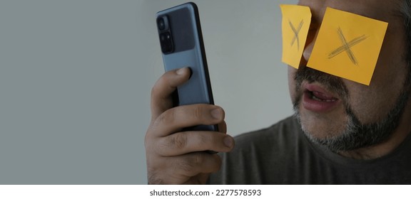 Stickers with painted crosses are glued to the eyes of a guy, a man is looking in a mobile cell phone with glued eyes, an isolated white background. shocked face - Shutterstock ID 2277578593