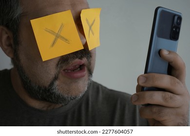 Stickers with painted crosses are glued to the eyes of a guy, a man is looking in a mobile cell phone with glued eyes, an isolated white background. shocked face - Shutterstock ID 2277604057
