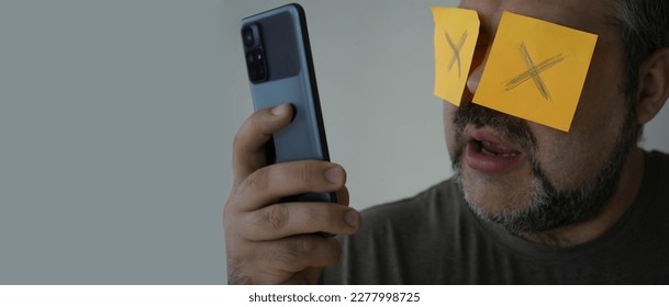 Stickers with painted crosses are glued to the eyes of a guy, a man is looking in a mobile cell phone with glued eyes, an isolated white background. shocked face - Shutterstock ID 2277998725