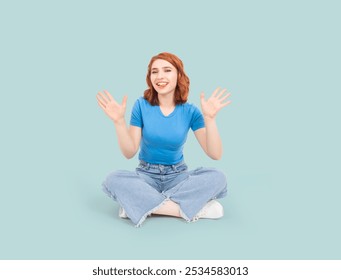 Sticking out her tongue, full body length beautiful red bob hair 20s woman sit floor sticking out her tongue hold up her arms show open palms looking camera. Funny young lady having great time.