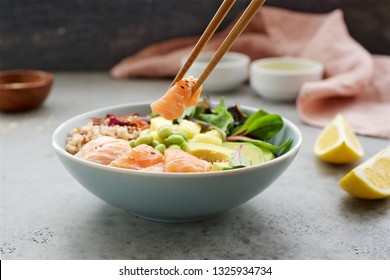 Sticks Holding A Piece Of Fresh Salmon. Salmon Poke Bowl With Salad, Avocado, Edamame Beans, Pineapple, Sesame And Brown Rice. Traditional Hawaiian Dish