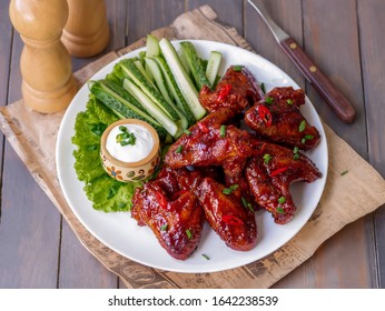 Sticky BBQ Chicken Wings With Dipping Sauce Served Hot With Cucumber Sticks On White Plate. Wooden Background, Selective Focus.