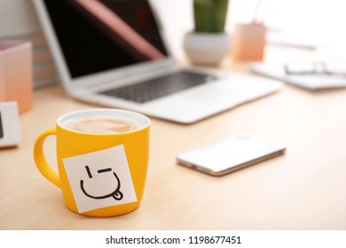 Sticky note with funny face attached to cup of coffee on office table. Space for text