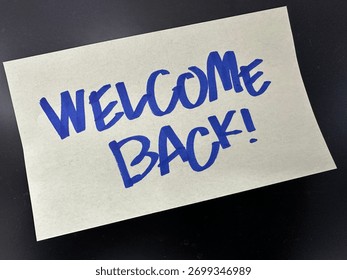 Sticky note with handwritten message “WELCOME BACK!” in bold blue ink, placed on a dark surface.