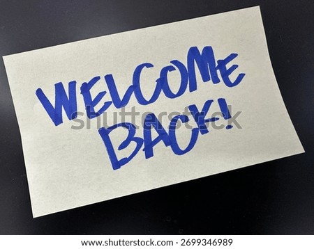 Sticky note with handwritten message “WELCOME BACK!” in bold blue ink, placed on a dark surface.