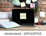 Sticky note with word SOS, laptop and stationery on wooden table at workplace