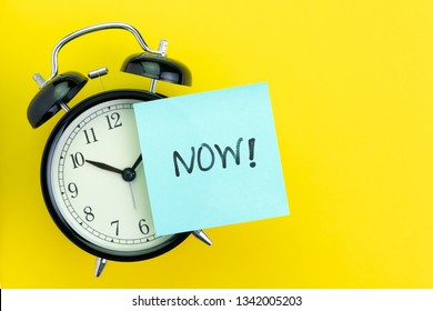 Sticky post with handwriting the word Now stick on alarm clock on solid yellow background with copy space using as stop procrastination, self discipline or motivation to complete task.
