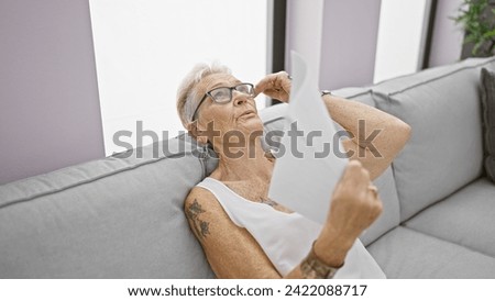 Stifling heat indoors, sweating, grey-haired senior woman, suffering from hot air, ingeniously uses documents as a handfan at home