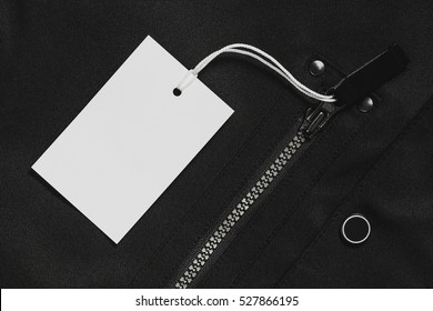 Still Life Blank Price Tag On A Dark Jacket With Zippers. Black And White Paper Cloth Label Mockup, Top View. 