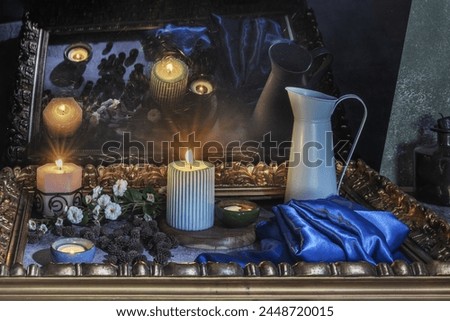 A still life with a blue satin fabric next to several lit candles, flowers surrounded by a golden frame and their reflection on a black surface