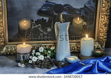 A still life with a blue satin fabric next to several lit candles, flowers and their reflection on a black surface