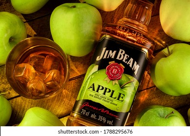 Still life with a bottle of Jim Beam Apple Bourbon (Apple liqueur from Kentucky) with a glass of cold whiskey on ice on a wooden brown background of boards, surrounded by juicy green apples.