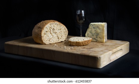 A Still Life Of A Bread Board Complete With A Slice Of Bread, Blue Cheese And A Glass Of Port Wine Set Against A Black Background