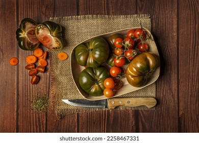 Still life with cherry tomatoes, large ripe tomatoes, carrot slices and a knife on burlap cloth and all on a dark wood panel - Shutterstock ID 2258467331