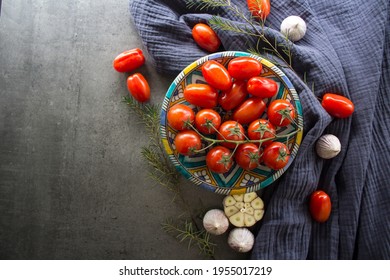 Still Life With Cherry Tomatoes On Ceramic Plate, Garlic Bulbs And Herbs On Navy Blue  Kitchen Towel. Gray Textured Background With Copy Space. 