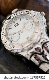 Still Life Close Up Detail View Of A Masculine Expensive Quality Waterproof Watch With Water Drops, Resting On A Bed Of Dark Natural Stones.  Object Detail With No People.