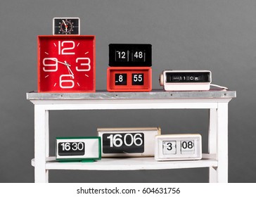 Still Life Of Collection Of Clocks In Variety Of Styles - Clock Radio, Flip Style, And Alarm Clocks - On White Table In Front Of Gray Background