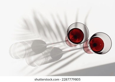 Still life with colored glass glasses of red wine, sunlight shadows and palm leaf shade, summer flat lay. Minimal style two stemmed wine glasses on white background, copy space, trend aesthetic photo  - Shutterstock ID 2287779805