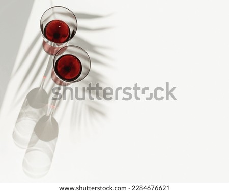 Still life with colored glass glasses of red wine, sunlight shadows and palm leaf shade, summer flat lay. Minimal style two stemmed wine glasses on white background, copy space, trend aesthetic photo 