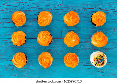 Still Life Of Colorful Cupcakes With Orange Twirled Icing Arranged In Neat Rows With One Lone White Cupcake With Sprinkles In A Conceptual Image Viewed From Above On Blue Wood