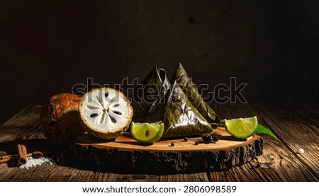 A still life composition featuring a halved cherimoya fruit, lime slices, and a pyramid-shaped, leaf-wrapped food item on a rustic wooden surface.