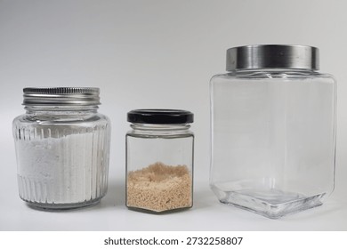 A still life composition featuring three distinct clear glass jars with metal lids, two of which contain white powder and brown granules, set against a clean white background.