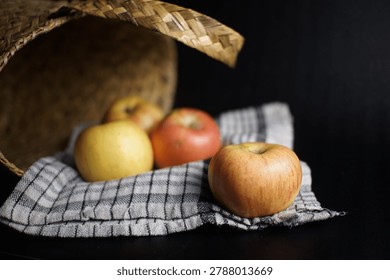 A still life composition featuring three vibrant apples, two red and one yellow, artfully arranged on a checkered cloth, with a rustic woven basket tilted in the background, all set against a dark, c.