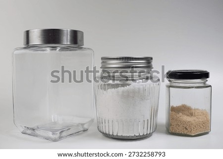 A still life composition featuring three clear glass jars of varying sizes, each with a metallic or black lid. One jar is empty, while the others contain white and brown powdered substances.