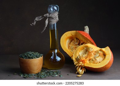 Still Life Consisting Of Sunflower Oil In A Decorative Bottle Of Two Halves Of Pumpkin And Lying Around Dried Shelled Fried Dark Pumpkin Seeds On A Table On A Dark Background