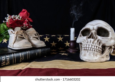 Still Life Depicting The Birth And Death Of A Nation With Baby Shoes On Bible And Skull And Smoking Candle On American Flag