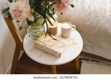 Still life details in home interior of living room. Open book with glasses, cup coffee and bouquet white pink peonies flowers. Read and rest. Cozy home
