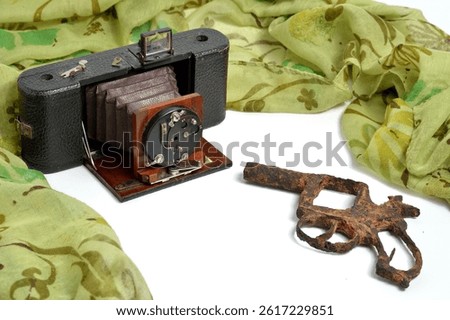 A still life featuring an antique bellows camera with its mechanism exposed, alongside a non-firing replica firearm, arranged with a light green patterned fabric on a white surface.