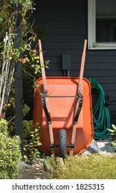 A Still Life Of Gardening Tools, Focusing On An Orange Wheel Barrow Leaning Against A House, With A Coiled Green Garden Hose, And Mulch.