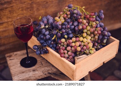 Still life with a glass of homemade red wine, wooden box with a variety of grapes, harvested in vineyards in a countryside, on a backless wooden stool on wood background. Viticulture, wine production - Shutterstock ID 2189071827