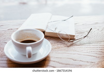 Still Life Image Of Cup Of Black Coffee On Saucer With Vintage Book And Eyeglasses On The Wooden Table Next To Big Coffee Shop Window.