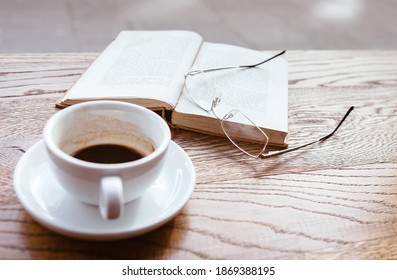 Still Life Image Of Cup Of Black Coffee On Saucer With Vintage Book And Eyeglasses On The Wooden Table Next To Big Coffee Shop Window.