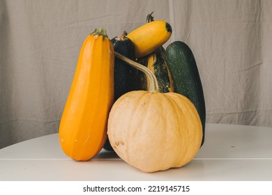 A Still Life Of One Pumpkin And Four Zucchini Stands On A White Table. The Still Life Is Lit By Natural Light From The Window. There Is A Linen Background. Autumn Harvest. October Harvest.