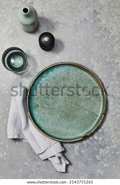 Still life\
photography of a green stoneware plate with napery, water glass,\
pepper grinder and a vase on a mottled grey background for food\
photography. Top view mock\
up.