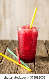 Still Life Profile Of Refreshing And Cool Frozen Red Fruit Slush Drink In Plastic Cup Served On Rustic Wooden Table With Collection Of Colorful Straws