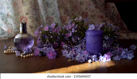 A Still Life Of Purple And Violet Phlox Flowers, A Perfume Transparent Bottle, A String Of Pearls And A Gradient Purple Candle On A Wooden Table In A Room With Curtains Behind In Shadow And Light