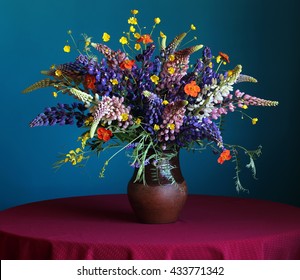 Still life with a summer bouquet of lupine and buttercups in the jug on the round table with a red tablecloth on a blue background.