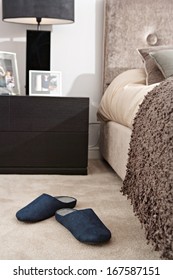 Still Life View Of An Elegant Master Bed In A Design Home Bedroom With Warm Colors And Fabrics, And A Pair Of Blue Slippers Resting On The Beige Carpet Floor. Home Interior View With No People.