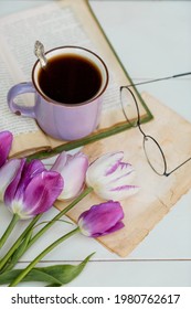 Still Life In Vintage Style. A Fancy Violet Coffee Mug, An Old Book, Tulips, And Glasses On A Wooden Table. 