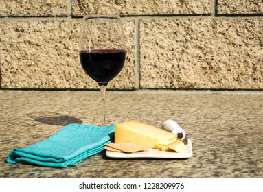 still life-glass of red wine with crackers, cheddar cheese, cheese knife on a white plate with a turquoise napkin outdoors against a stone wall - Powered by Shutterstock