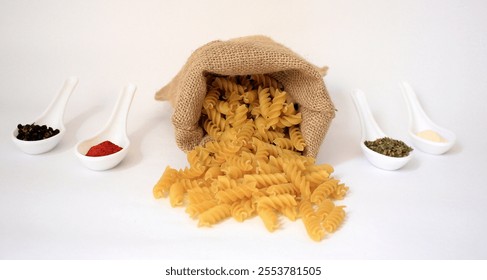 A still-life composition featuring a burlap jute sack positioned in the center of the frame, filled with uncooked fusilli pasta. On either side of the sack, several ceramic spoons with spices  - Powered by Shutterstock