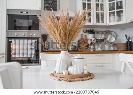 Still-life. Dried pampas grass in a vase, white ceramic pumpkins, a teapot and pumpkin-shaped candles on a white table in the interior of a Scandinavian-style home kitchen. Cozy autumn concept.