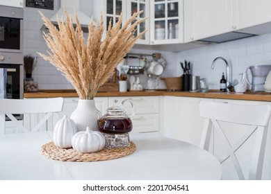 Still-life. Pampas dried grass in a vase, white ceramic pumpkins and a teapot on a white table in the interior of a Scandinavian-style home kitchen. Cozy, stylish autumn concept.