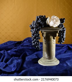 Still-life Of A Pedistal With Concord Grapes With A White Rose On Royal Blue Stain And Gold Colored Background.