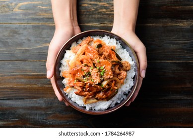 Stir fried kimchi with pork on cooked rice in a bowl holding by hand on wooden background, Korean food, Top view